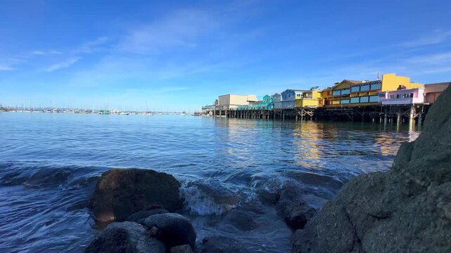 Low Angle View of Old Fishermen's Wharf from Beach Rocks with Sailing Boats on the Horizon and Small Waves 4K.