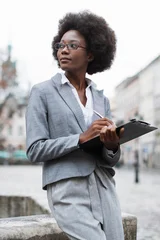 Selbstklebende Fototapeten Altes Krankenhaus Beelitz A professional African American woman in a suit and glasses is thoughtfully writing notes on a clipboard while standing outside in an urban setting  © sofiko14