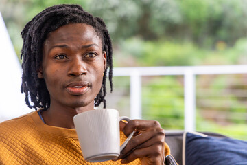 African American man sitting on covered balcony holding white ribbed mug wearing mustard sweatshirt