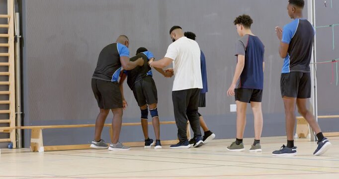 Diverse male athletes and coach guiding injured athlete limping toward bench in gym after stumbling