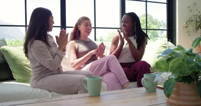 Diverse female friends embracing after comforting hug, laughing and talking on sofa with teal mugs