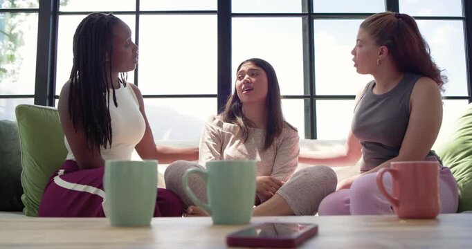 Diverse female friends hugging center woman clutching chest, draping blanket on sofa at home, phone