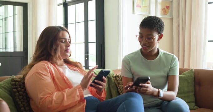 Diverse women on sofa, left woman scrolling phone, sparking right woman checking phone, laughing