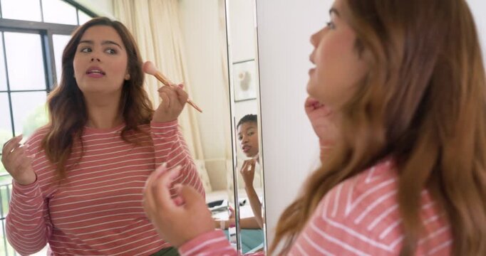 Female friends, standing woman applying bronzer with face brush at bedroom mirror, getting ready