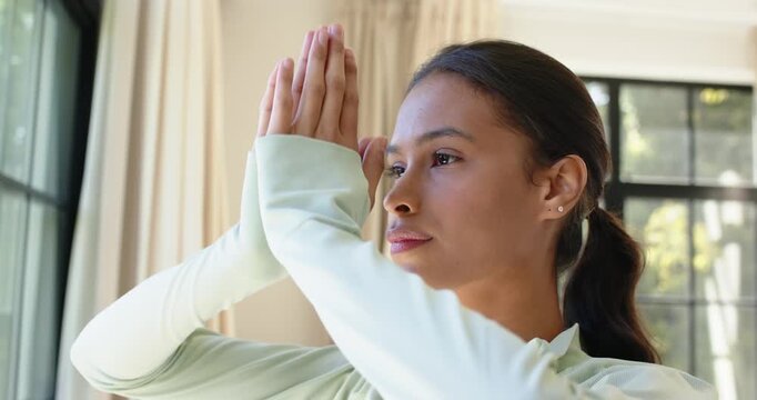 Adult African woman lifting arms, pressing palms into prayer near window in green top, meditating