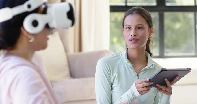 Diverse female guide tapping tablet and speaking, coaching VR user wearing white headset at home
