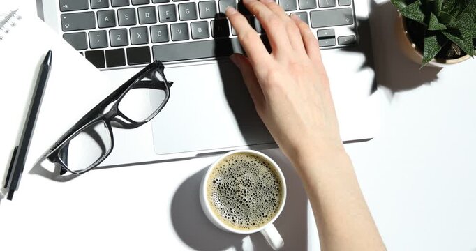 Woman putting cup of coffee down and working on laptop at white desk, top view