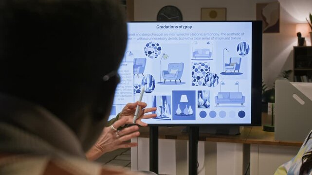 Senior female interior designer introducing colleagues to modern trends in gray palettes pointing at smart display in office meeting room