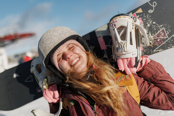 Girl enjoying winter fun while holding snowboard on her shoulders at a ski resort during a sunny day in the mountains with clear blue sky above