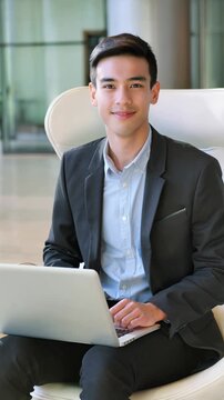 Happy young businessman giving a thumbs-up while working on a laptop in a modern office lobby, vertical shot