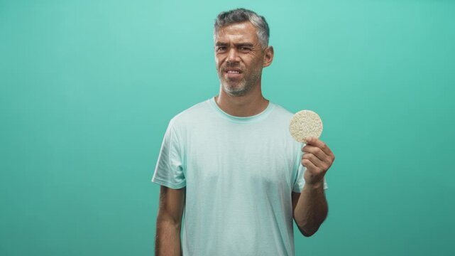 Man holding a round corn pastry in his right hand, eyes closed and slight smile, posed against studio wall; satisfaction.