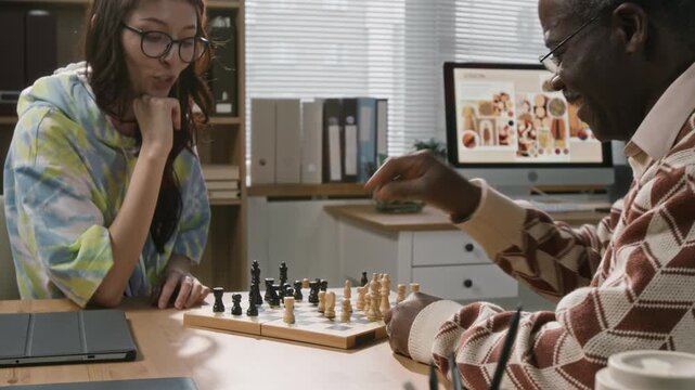Cheerful young adult woman moving piece on chessboard competing with senior Black male colleague in intellectual game during work break at office