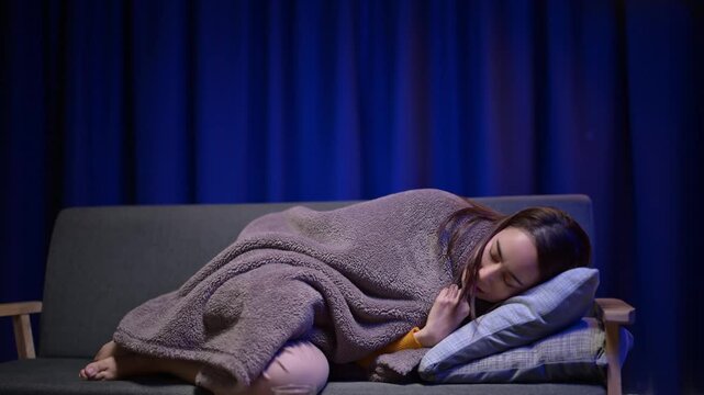 A woman is sitting on a couch wrapped in a blanket. She is wearing a yellow shirt and she is cold