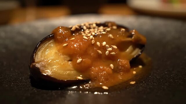 Close-up of Nasu Dengaku (Grilled Miso Eggplant) served on a dark plate, garnished with sesame seeds in a gourmet restaurant setting.