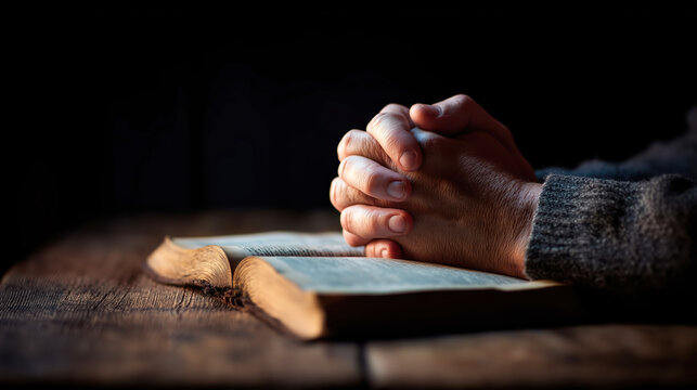 Two hands are joined together in prayer on an open Holly bibble laid on a wooden table. Soft light falls on the scene, creating a thoughtful atmosphere during a reflective moment.