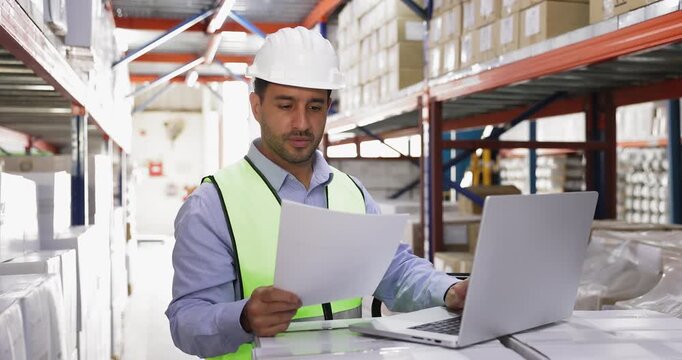 Male Hispanic warehouse manager man in hard hat working at laptop, checking waybill, consulting electronic consignment, using logistic app on computer in storage space
