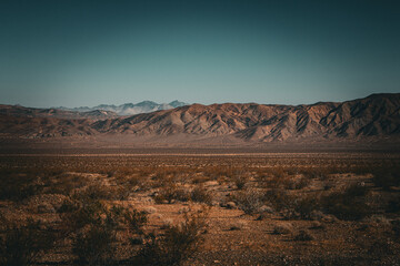 Naklejka premium A vast desert landscape with dry earth sparse bushes and distant rugged mountains under a clear blue sky creates a sense of solitude and natural beauty