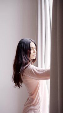 Attractive Young Female Opening Window Curtains to Let Bright Morning Sunlight Enter Room. Alone Adult Girl Looking Outside With Hope. One Beautiful Woman Starting Her Day in Cozy Modern Residence 4k