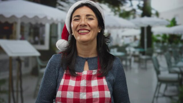 Woman in red santa hat and checkered apron extends hand for handshake at outdoor terrace near building facade; hospitality.