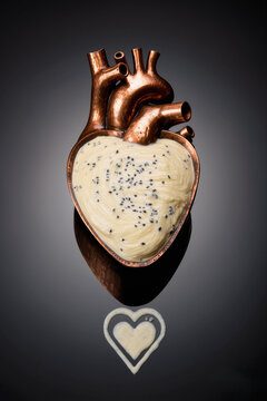 A copper heart shaped anatomical model filled with white cream and black sesame seeds on dark reflective surface, romantic dessert concept.