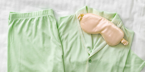 Green Pajama Set with peach color sleep mask on white sheet. Top view trend pale green woman home sleepwear from soft cotton on bedclothes. Cozy pyjamas of shirt, shorts for comfort rest, flat lay