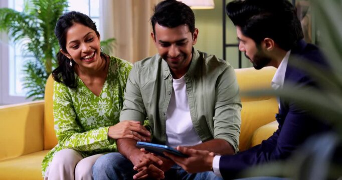 Indian couple discussing with agent at home with tablet computer during financial investment meeting with bank representative reviewing documents while sitting on sofa in cozy modern living room inter