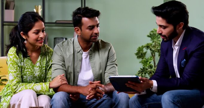 Indian couple discussing with agent at home with tablet computer during financial investment meeting with bank representative reviewing documents while sitting on sofa in cozy modern living room inter