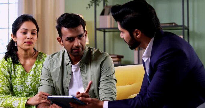 Indian couple discussing with agent at home with tablet computer during financial investment meeting with bank representative reviewing documents while sitting on sofa in cozy modern living room inter