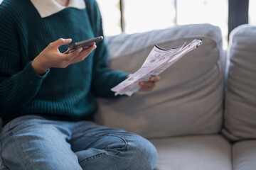 Asian woman holding paper document calculating money savings paying bills in mobile application using smart phone payment online with paying household bills tax insurance managing budget.