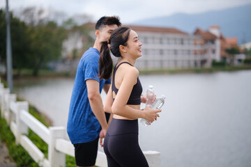 Naklejka premium Active couple taking a break after outdoor running, holding water bottles while enjoying a peaceful lakeside view. Healthy lifestyle concept.