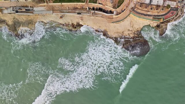 Sideways aerial view of a coastal construction site at the St Thomas Bay in Marsaskala, Malta, with waves crashing against the rocky Mediterranean shore, showing the interplay of urban development.
