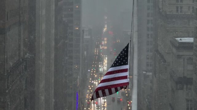 Aerial view of American flag waving above downtown street during winter storm, with falling snow, fog and glowing yellow headlights. Moody urban atmosphere in American town. Close up.