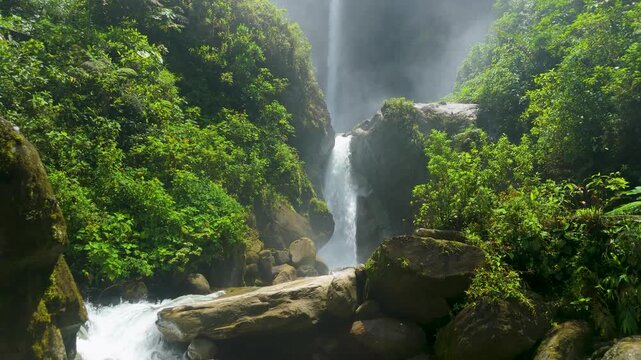 Immersive low-altitude drone flight skims the river and jungle canopy, approaching the base of the majestic Cascada El Rocio Machay waterfall in the lush tropical mountains of Ecuador