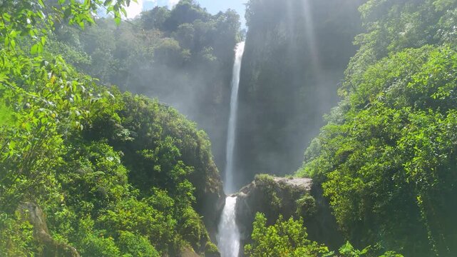 Low angle aerial glide moves forward and tilts up to reveal the majestic Cascada El Rocio in Machay, cascading through misty cliffs and vibrant green rainforest near Banos, Ecuador.