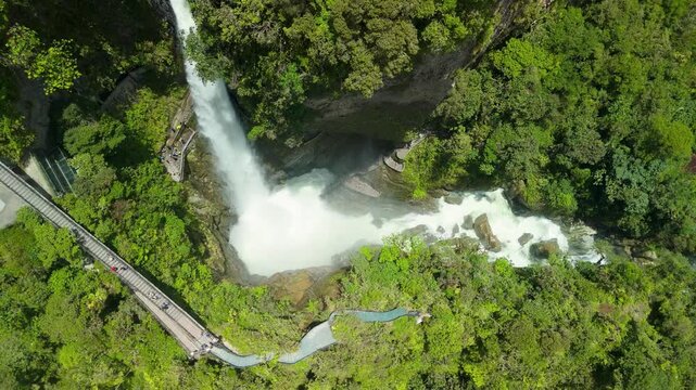 Aerial top-down parallax shot captures the massive Pailon del Diablo waterfall crashing into the canyon alongside lush jungle and people navigating the famous zigzagging stone stairs in Banos Ecuador