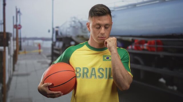 Man holding basketball with hand, cheek resting on fist while wearing brasil jersey with flag patch and dockside vehicle visible on street; pensive reflection.