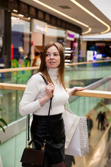 Fototapeta premium An adult woman stands inside a contemporary mall, looking content while holding shopping bags. She gazes around, showcasing a relaxed lifestyle during her shopping trip in a bustling environment.