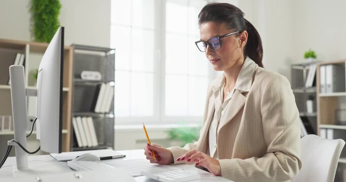 Accountant auditor businesswoman with glasses doing finance planning. She types on a computer in an office, reviewing paperwork with audit analysis. Clear concept of precise financial oversight.