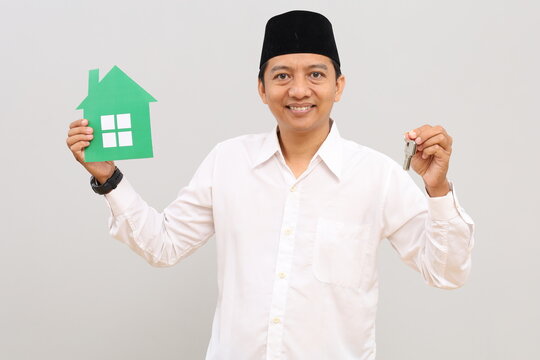 Asian Muslim man holds a small paper house image and a house key, posing in front of a gray background wall with empty space for promotional content or design.