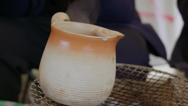 A rustic ceramic pitcher with a two-tone glaze&mdash;pale off-white at the bottom. Steam rises from metal grill's opening. Grill holds a hot drink. Cozy, casual outdoor scene.