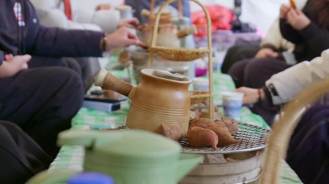 A group of people gather around a low table. Warm, casual outdoor activity. Ceramic pot sits on metal grill, surrounded by roasted sweet potatoes. Cozy, convivial outdoor atmosphere.