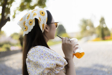 Relaxed asian woman enjoying refreshing summer drink in sunny outdoor park. fashionable young female with headscarf and sunglasses enjoying peaceful lifestyle