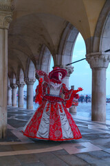 Full-length portrait of Venetian carnival performer wearing red and silver ornate dress with decorative fan under stone arches in Venice