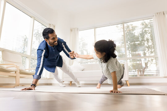Young sportive Indian father and little son fist bumping, making gesture of encouragement and praise, performing one-armed push-up, plank exercise in sunny living room. Teamwork, family fun, support