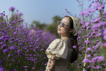 Happy young Asian woman in sunglasses enjoying serene moment in beautiful purple verbena flower field. beautiful lady wears dress outdoors under sunny sky