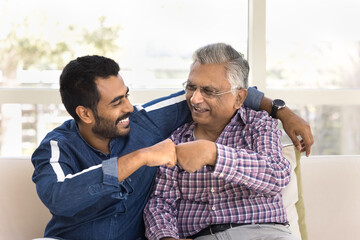 Happy young Indian man hugging and fist bumping older grandpa or great-grandfather seated together...