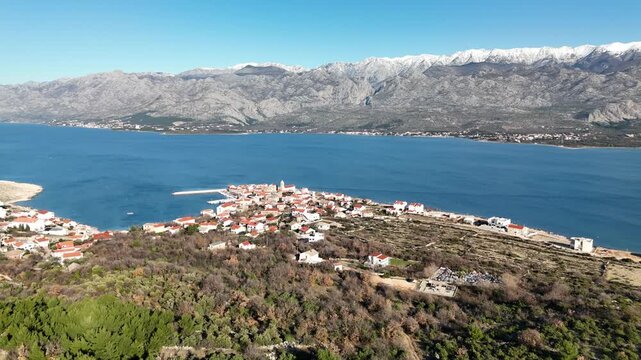 Drone flying from green hills toward Vinjerac, revealing Adriatic coastline and massive Velebit mountains before approaching the coastal town on a sunny winter day in Dalmatia, Croatia.