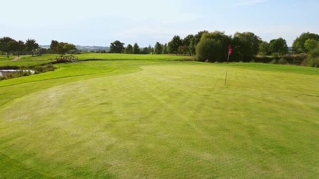 Low drone flying over a green fairway towards the red flag on a putting green of a golf course. Aerial forward, copy space