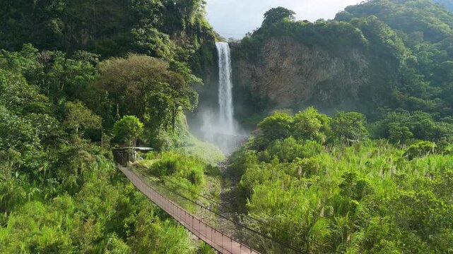 Drone flight approaching magnificent Manto de la Novia waterfall and long pedestrian suspension bridge over lush green tropical valley in Banos Ecuador highlands