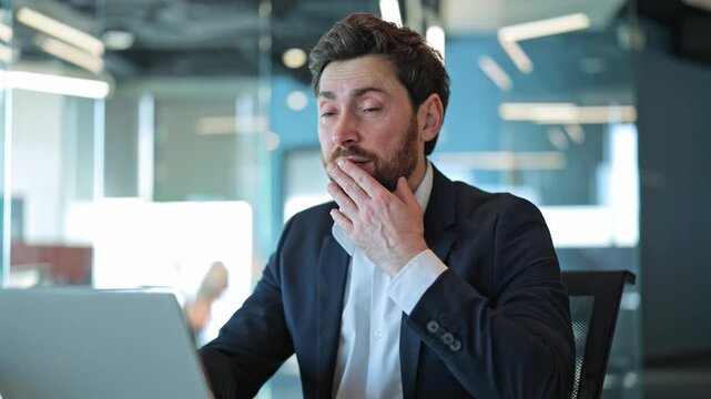 Tired professional working on laptop. Yawning and eventually dozing off at modern office desk, feeling overwhelmed. Represents burnout, fatigue, and lack of sleep from demanding work.